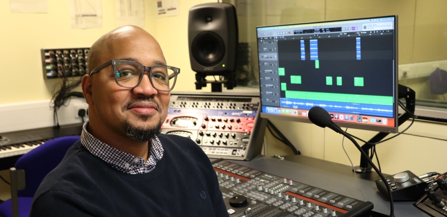 An African male wearing glasses and a navy jumper over a shirt sits in a music recording studio in front of a monitor and sound desk.